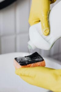 A person wearing yellow gloves pouring cleaning liquid onto a sponge, showing the use of proper cleaning tools and methods.