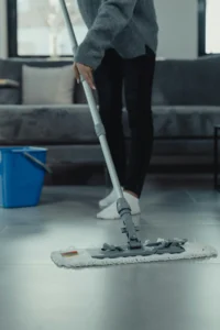 A cleaner from Gloria Cleaning is mopping a tiled floor in a cozy living room, with a blue bucket nearby, showcasing their professional service and the benefits of hiring their team